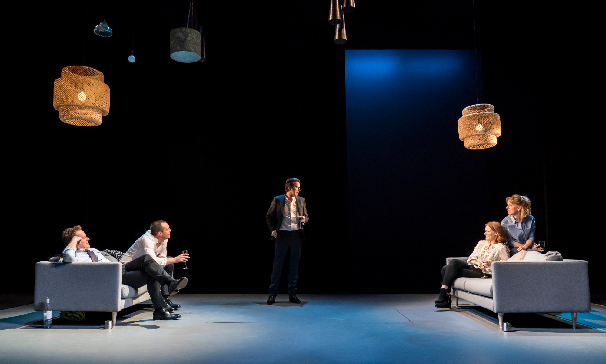 Two sofas are on stage on the far left and far right. Two actors are sitting on each. In the middle is a male who is looking at someone on the right sofa.