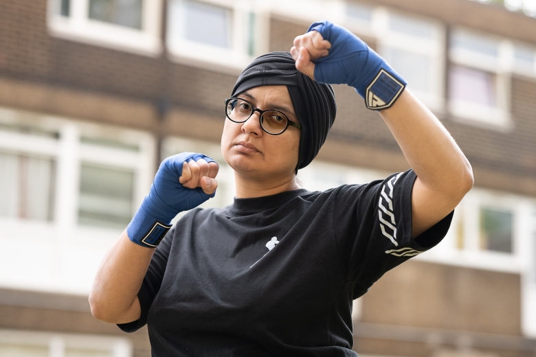 A woman wears a black headscarf, glasses and a black shirt. She has wraparound boxing gloves around her knuckles and wrists, which are raised in a fighting pose. Behind her are a block of flats.