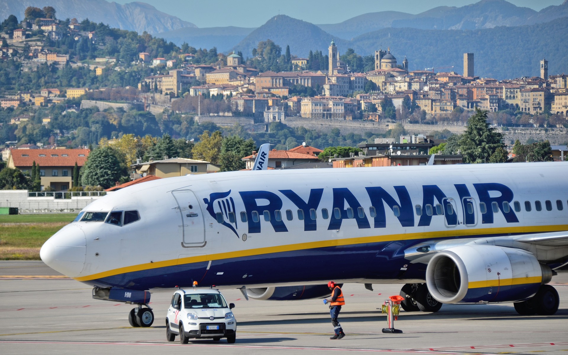 A side-on view of a Ryanair plane on the runway. Behind the plane is a city in the distance.