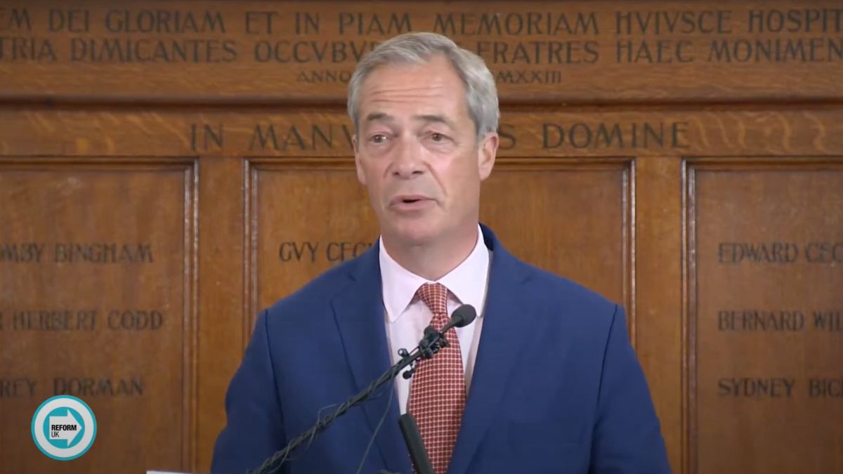 Nigel Farage in a blue suit, speaking in front of a wooden wall with lettering on it.